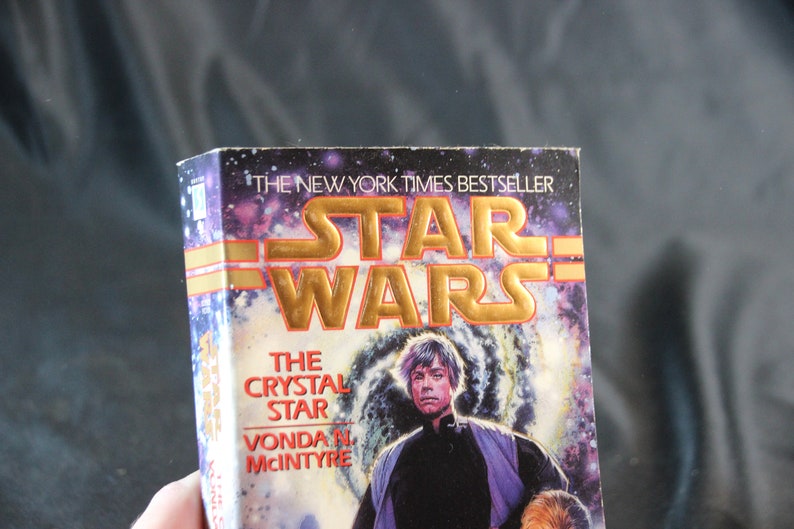 May include: A paperback book titled "Star Wars: The Crystal Star" by Vonda N. McIntyre. The cover features a space scene with a portrait of a man. The title is in gold lettering, and the book is a New York Times bestseller.
