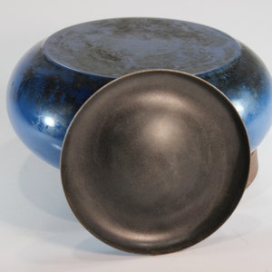 May include: A blue ceramic bowl with a dark, textured interior and a matching lid. The bowl has a rounded shape and a glossy finish. The lid is a dark gray color. The bowl and lid are likely handmade.