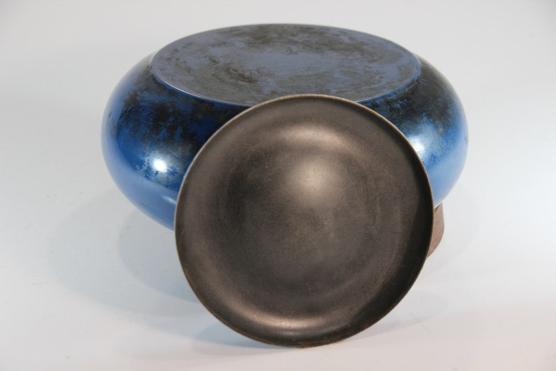 May include: A blue ceramic bowl with a dark, textured interior and a matching lid. The bowl has a rounded shape and a glossy finish. The lid is a dark gray color. The bowl and lid are likely handmade.