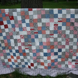 May include: A handmade quilt with a patchwork design. The quilt features a grid of squares in red, white, blue, and gray fabrics. The border is a patterned fabric in shades of gray and white. The quilt is draped over a wooden fence.