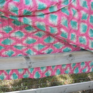 May include: A pink and teal patterned fabric draped over a wooden fence. The fabric features a repeating design of teal diamond shapes on a pink background, with a grid pattern overlay. The fence is made of weathered wood and wire.