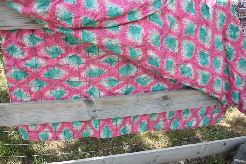 May include: A pink and teal patterned fabric draped over a wooden fence. The fabric features a repeating design of teal diamond shapes on a pink background, with a grid pattern overlay. The fence is made of weathered wood and wire.