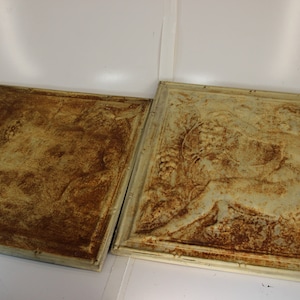 May include: Two square, antique-style wall panels with a distressed, textured finish. One panel features a detailed relief design of a figure and foliage, while the other has a simpler, abstract pattern. The panels have a rustic, aged appearance.