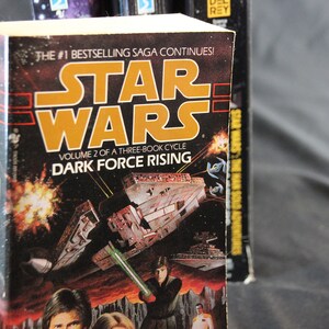 May include: A hardcover book titled "Star Wars: Dark Force Rising." The cover features a space battle scene with a lightsaber and the Star Wars logo in orange. The text reads "Volume 2 of a Three-Book Cycle."