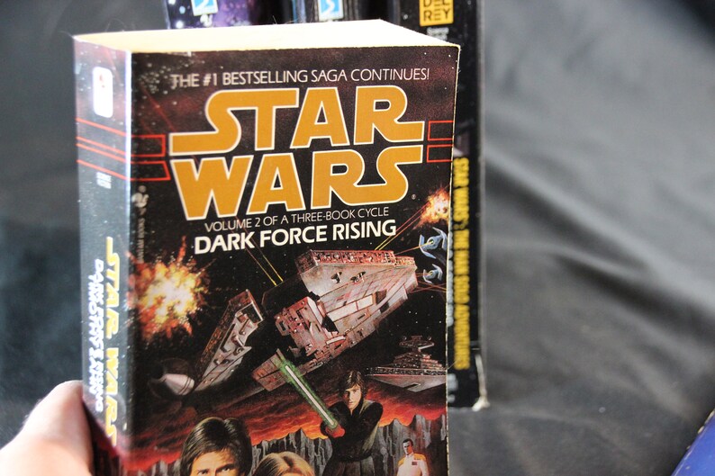 May include: A hardcover book titled "Star Wars: Dark Force Rising." The cover features a space battle scene with a lightsaber and the Star Wars logo in orange. The text reads "Volume 2 of a Three-Book Cycle."