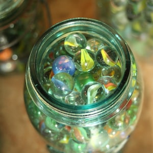 May include: A glass jar filled with colourful marbles. The marbles are a mix of blue, green, yellow, and white. The jar is sitting on a wooden surface.