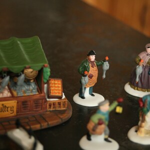 May include: A collection of miniature ceramic figurines depicting a winter market scene. The figurines include a poultry stand with a green awning, vendors holding poultry, and shoppers. The word "poultry" is visible on the stand.