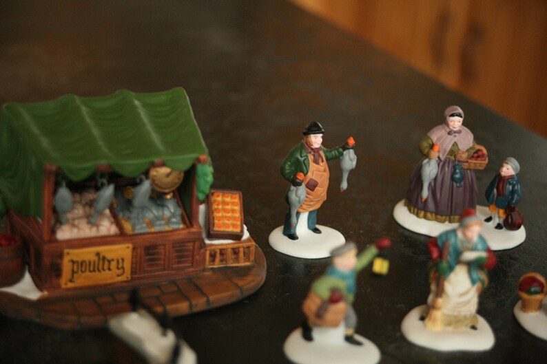 May include: A collection of miniature ceramic figurines depicting a winter market scene. The figurines include a poultry stand with a green awning, vendors holding poultry, and shoppers. The word "poultry" is visible on the stand.