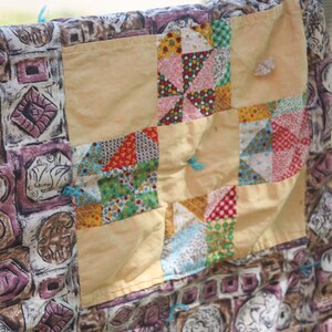 May include: A handmade quilt with a patchwork design. The quilt features a variety of patterned fabrics in shades of beige, brown, pink, red, and blue. The quilt is hanging on a metal fence.
