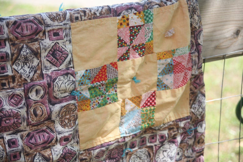 May include: A handmade quilt with a patchwork design. The quilt features a variety of patterned fabrics in shades of beige, brown, pink, red, and blue. The quilt is hanging on a metal fence.