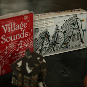 May include: A red "Village Sounds" book with musical notes and snowflakes, next to a box with a black and white illustration of a snowy village scene with string lights. A small house figurine is in the foreground.