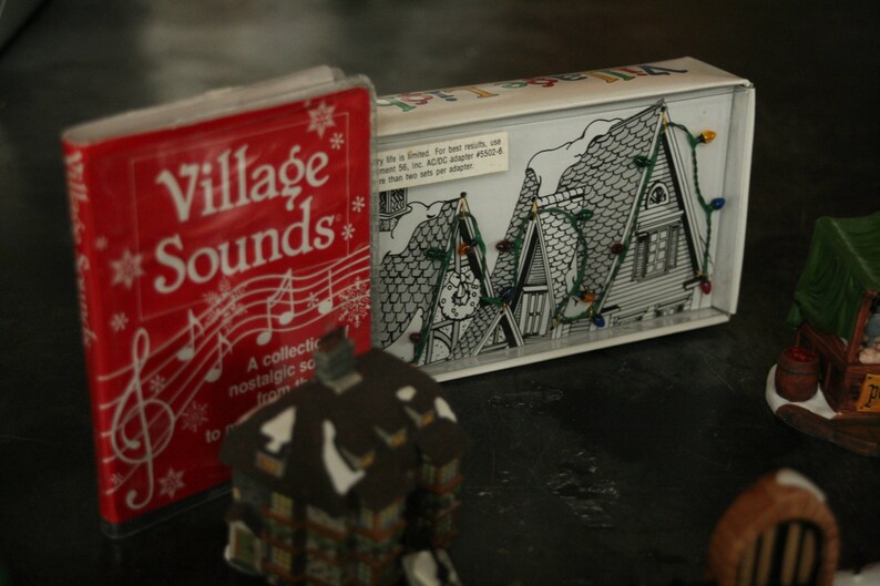 May include: A red "Village Sounds" book with musical notes and snowflakes, next to a box with a black and white illustration of a snowy village scene with string lights. A small house figurine is in the foreground.