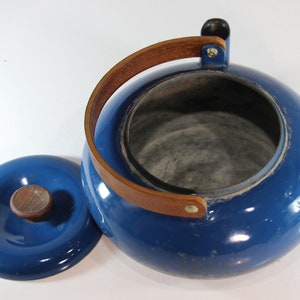 May include: A blue enamel teapot with a wooden handle and lid. The teapot has a round body and a wooden handle that arches over the top. The lid has a wooden knob. The teapot measures approximately 8 inches in diameter.