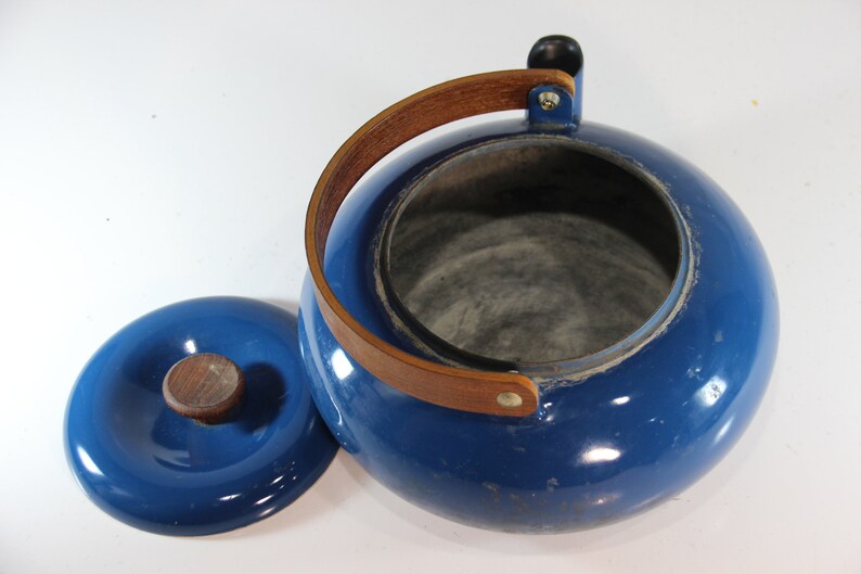 May include: A blue enamel teapot with a wooden handle and lid. The teapot has a round body and a wooden handle that arches over the top. The lid has a wooden knob. The teapot measures approximately 8 inches in diameter.
