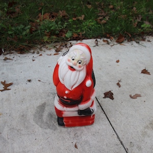May include: A vintage Santa Claus figurine, primarily red and white, with a black bag over his shoulder. The figurine has a smiling face and is standing on a red and black base. The figure is made of ceramic or a similar material.
