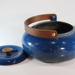 May include: A blue enamel kettle with a wooden handle and lid. The kettle has a round shape and a wooden handle that arches over the top. The lid has a wooden knob. The kettle is approximately 8 inches in diameter.