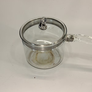 May include: A clear glass saucepan with a matching lid and a clear handle. The saucepan has a silver-colored rim and a clear handle. The lid has a silver-colored handle. The saucepan is designed for cooking.