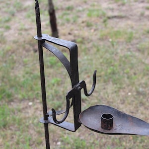 May include: A vintage-style wrought iron candle holder. It features a long vertical rod, a curved arm, and a small, shallow dish for the candle. The metal has a dark, aged finish. The design is simple and functional, with a rustic aesthetic.