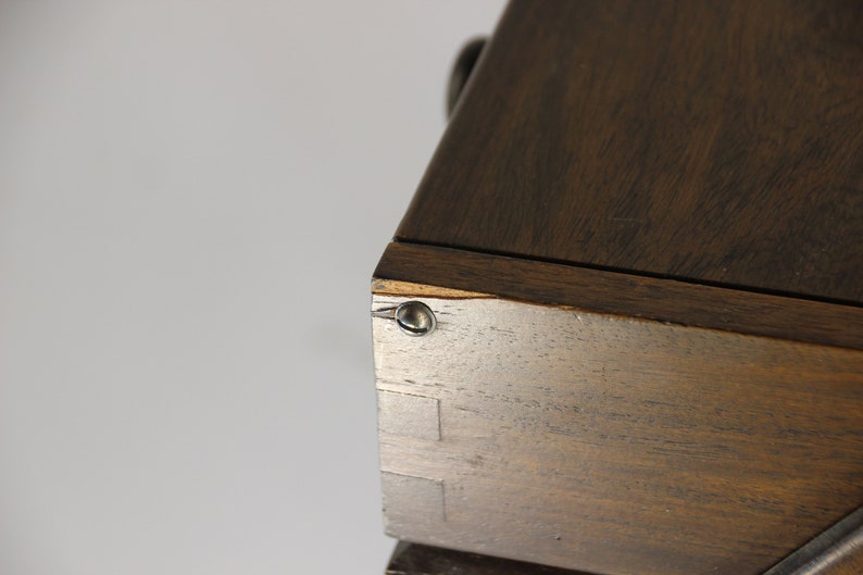 Puede incluir: Primer plano de una esquina de mueble de madera. La madera es de color marr&oacute;n oscuro con vetas visibles. La esquina est&aacute; unida con ensambles de cola de milano y asegurada con un remache met&aacute;lico. La pieza parece ser parte de un armario o cofre m&aacute;s grande.