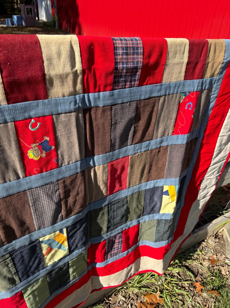 May include: A patchwork quilt with a variety of fabrics, including corduroy and patterned textiles. The quilt features a red border and blue accents, with a design that includes a horseshoe and a cowboy.