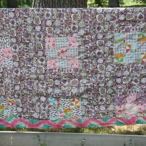 May include: A handmade quilt with a patchwork design. The quilt features squares in shades of pink, purple, and brown, with a decorative border of pink and teal zigzags. The quilt is draped over a wooden fence.