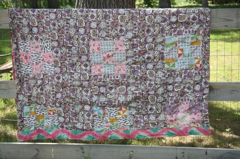 May include: A handmade quilt with a patchwork design. The quilt features squares in shades of pink, purple, and brown, with a decorative border of pink and teal zigzags. The quilt is draped over a wooden fence.