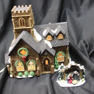 May include: A detailed Christmas village scene featuring a church with a tall steeple and a nativity scene. The church has a brown brick facade, a dark grey roof with snow accents, and lighted windows. The nativity scene includes figures of Mary, Joseph, and baby Jesus.