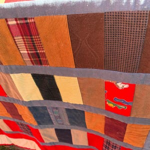 May include: A patchwork quilt with a variety of fabrics in shades of brown, orange, red, and blue. The quilt features corduroy, plaid, and patterned fabrics, with a red and white striped border.