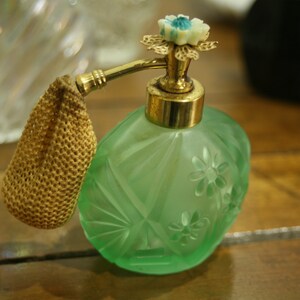 May include: A vintage green glass perfume bottle with a gold-colored atomizer and a decorative floral top. The bottle has a textured surface with diamond and flower patterns. A small, gold-colored mesh pouch is attached.