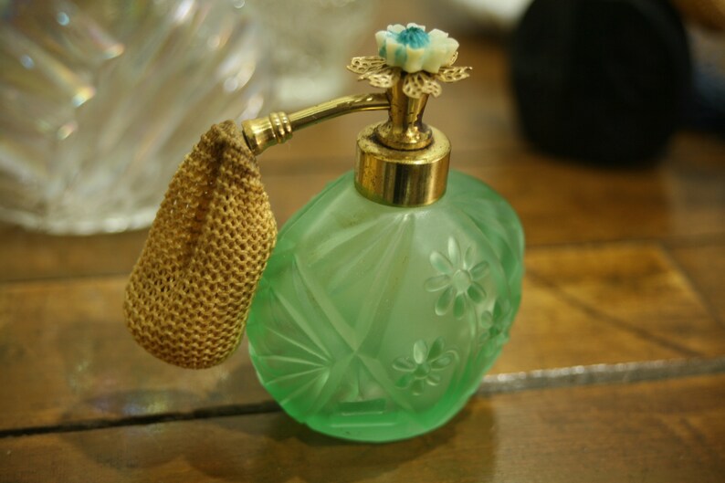 May include: A vintage green glass perfume bottle with a gold-colored atomizer and a decorative floral top. The bottle has a textured surface with diamond and flower patterns. A small, gold-colored mesh pouch is attached.
