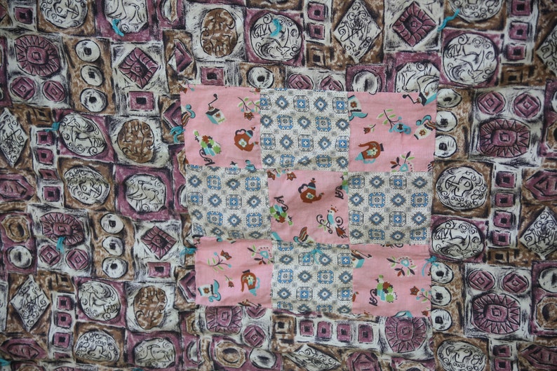 May include: A patchwork textile featuring a mix of patterns. The background has a repeating design of geometric shapes in brown, purple, and white. A pink square with floral and teapot motifs is sewn on top.