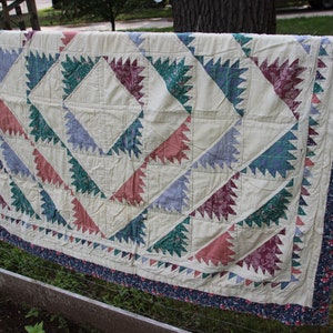 May include: A handmade quilt with a cream-colored background and geometric patterns in shades of red, green, and blue. The quilt is bordered with a floral print and is draped over a wooden fence.