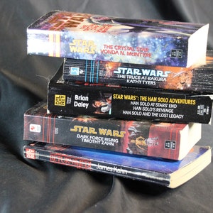 May include: A stack of Star Wars novels, featuring titles like "The Crystal Star" and "Dark Force Rising." The books have colorful covers with space-themed artwork and text. The books are stacked on top of each other.