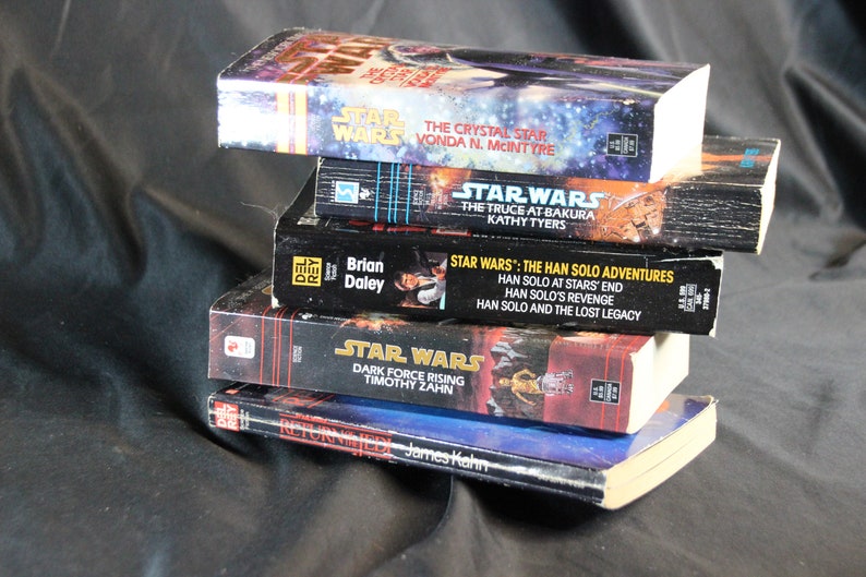 May include: A stack of Star Wars novels, featuring titles like "The Crystal Star" and "Dark Force Rising." The books have colorful covers with space-themed artwork and text. The books are stacked on top of each other.
