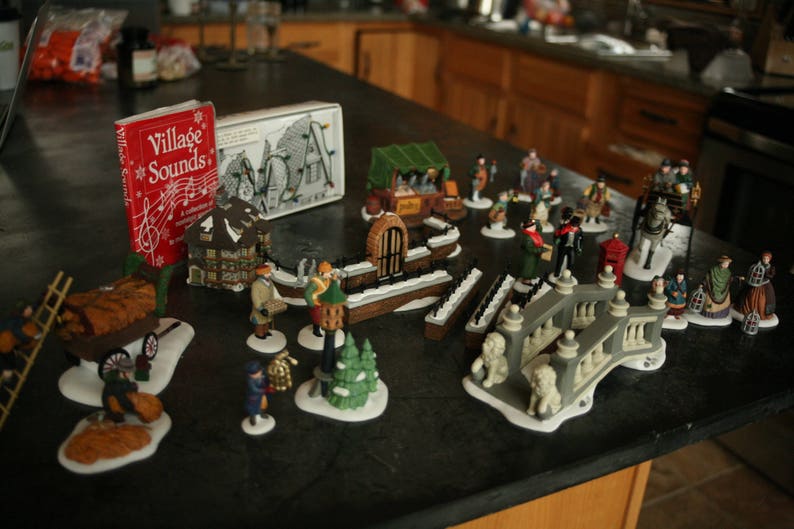 May include: A collection of miniature Christmas village figurines and accessories, including buildings, people, and decorative elements. A red box with the text "Village Sounds" is also present.