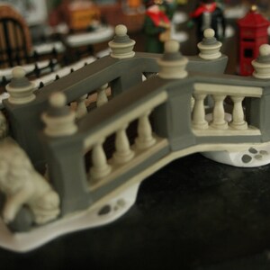 May include: A miniature gray and cream-colored decorative bridge with balustrades and ornate details. A small statue of a lion is at the base of the bridge. The bridge is part of a Christmas village scene.