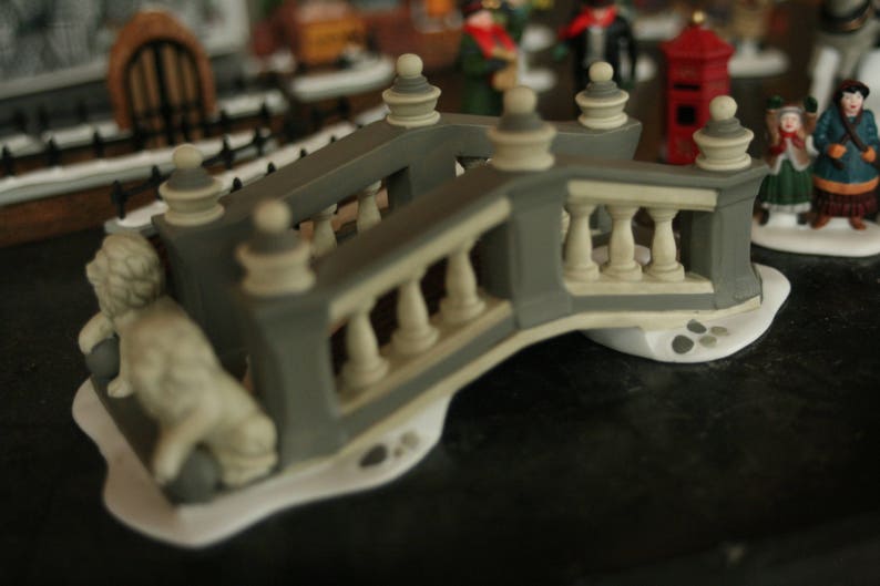 May include: A miniature gray and cream-colored decorative bridge with balustrades and ornate details. A small statue of a lion is at the base of the bridge. The bridge is part of a Christmas village scene.