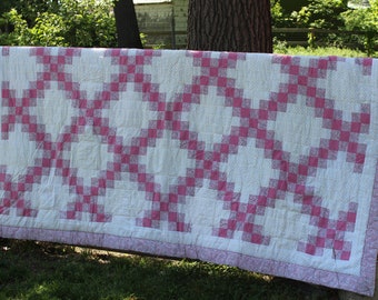 May include: A handmade quilt with a white background and a pink and white checkerboard pattern. The quilt is hanging on a wooden fence outdoors. The border is a solid pink color.