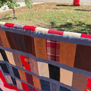 May include: A colorful patchwork quilt with a variety of fabric patterns and textures. The quilt features red, blue, brown, and cream-colored fabrics, arranged in vertical stripes. The edges are trimmed with a blue border and red and white stripes.