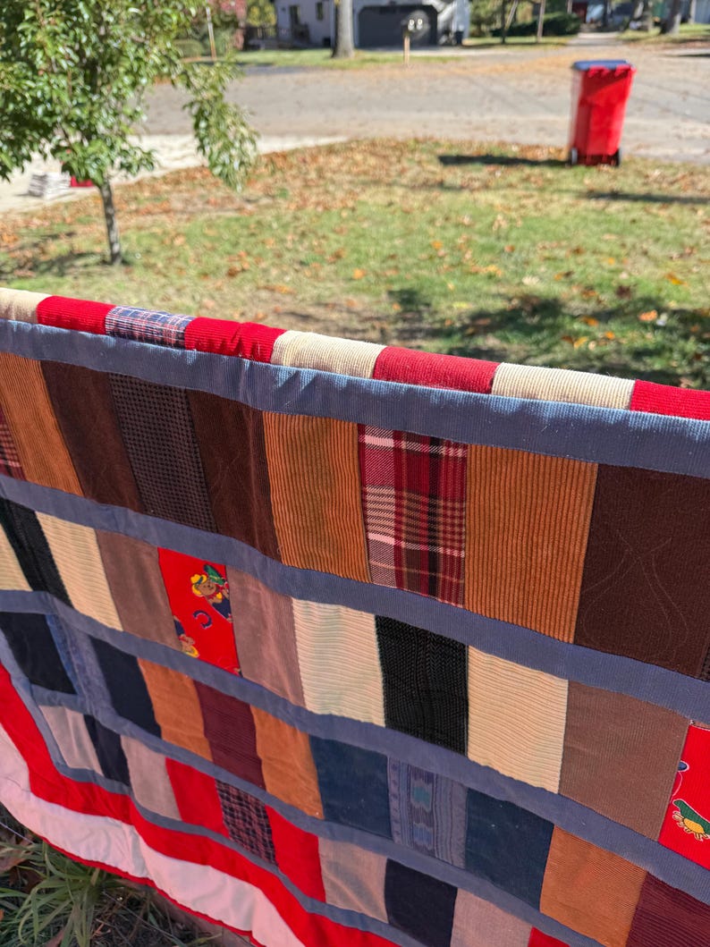 May include: A colorful patchwork quilt with a variety of fabric patterns and textures. The quilt features red, blue, brown, and cream-colored fabrics, arranged in vertical stripes. The edges are trimmed with a blue border and red and white stripes.