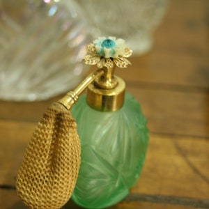 May include: A vintage green glass perfume bottle with a gold-colored atomizer and a decorative flower-shaped top. The bottle has an etched floral design and a woven beige bulb. Other bottles are in the background.