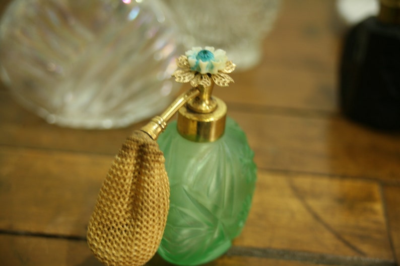 May include: A vintage green glass perfume bottle with a gold-colored atomizer and a decorative flower-shaped top. The bottle has an etched floral design and a woven beige bulb. Other bottles are in the background.