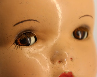 May include: Close-up of a vintage doll's face. The doll has brown eyes with detailed eyelashes, painted eyebrows, and a red painted mouth. The skin tone is a pale peach color, with visible age-related wear and tear.