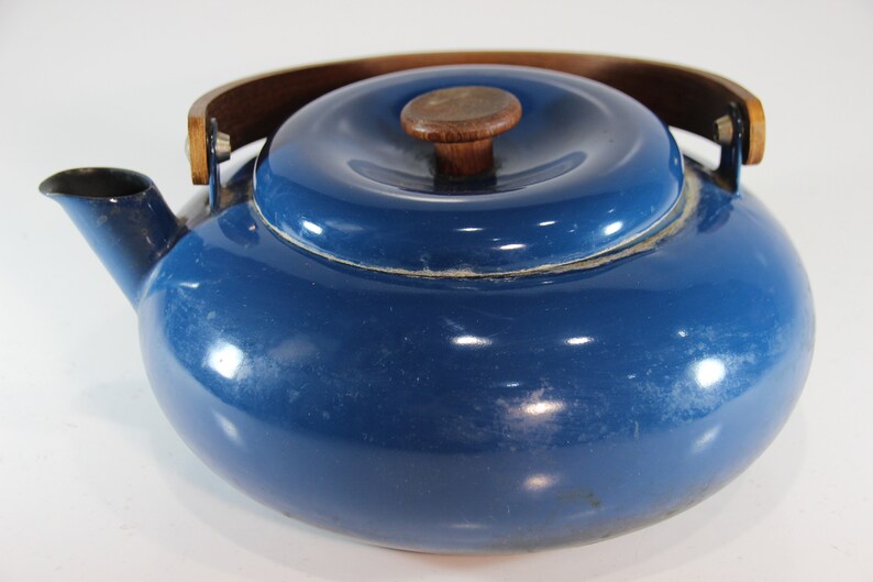 May include: A vintage blue enamel teapot with a wooden handle and lid knob. The teapot has a rounded body, a spout, and a lid. The wooden handle arches over the top, and the lid has a small wooden knob. The teapot is a classic kitchenware item.