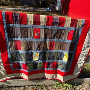 May include: A handmade quilt with a patchwork design. The quilt features a variety of fabrics in red, blue, brown, and beige. The quilt is hanging on a wooden fence outdoors.