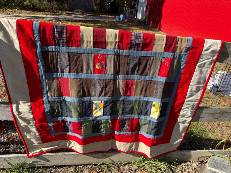 May include: A handmade quilt with a patchwork design. The quilt features a variety of fabrics in red, blue, brown, and beige. The quilt is hanging on a wooden fence outdoors.