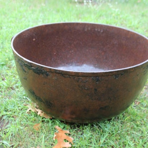 May include: A large, antique cast iron cauldron with a rounded shape and a dark, rusty brown color. The cauldron has two small handles and sits on a bed of green grass. The interior appears to be smooth and reflective.