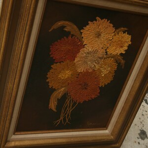 May include: A framed floral painting featuring a bouquet of orange, red, and yellow flowers against a dark brown background. The flowers are painted with textured brushstrokes, and the frame is gold-colored.