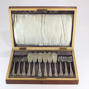 Puede incluir: Un juego de cubiertos de madera con tapa con bisagras, que contiene tenedores y cuchillos de mantequilla. La cubertería está dispuesta en un interior forrado de terciopelo, con una tela de color claro en la parte superior de la caja. La caja se cierra con un pequeño pestillo.