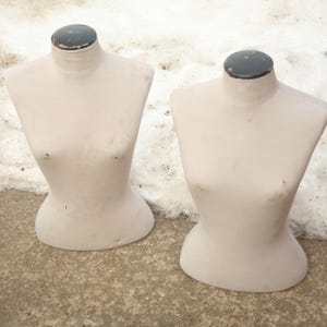 May include: Two vintage dressmaker's mannequins. The torso forms are a pale pink color with a black cap on the neck. The mannequins are standing on a concrete surface with snow in the background. The forms are used for displaying clothing.